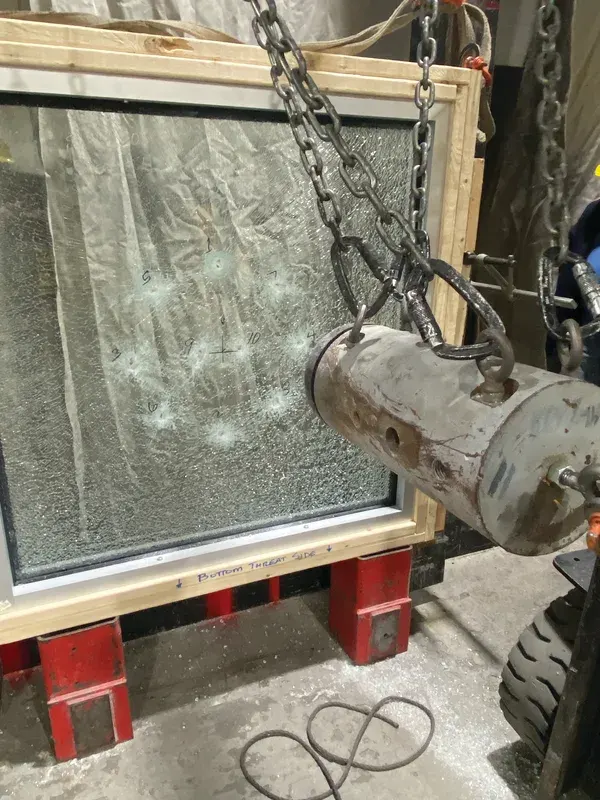 A laminated pane in a wooden frame on red stands, held against a chained steel impactor, with numbered impact marks on the glass and BOTTOM THREAT SIDE written on tape along the sill.