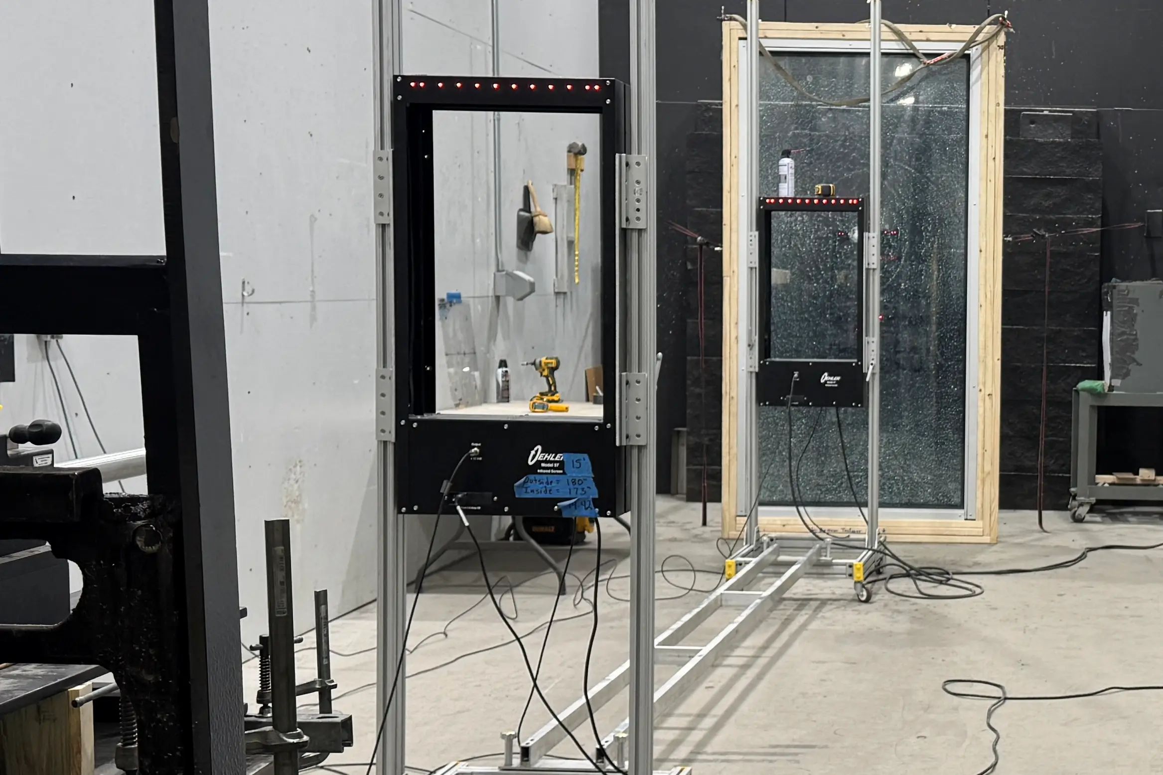 RiotLite's forced entry test bay: two glazing test frames on rails, Oehler chronograph screens with lit red rows, a blue tape range card, and a crazed laminated pane held in a wooden frame.