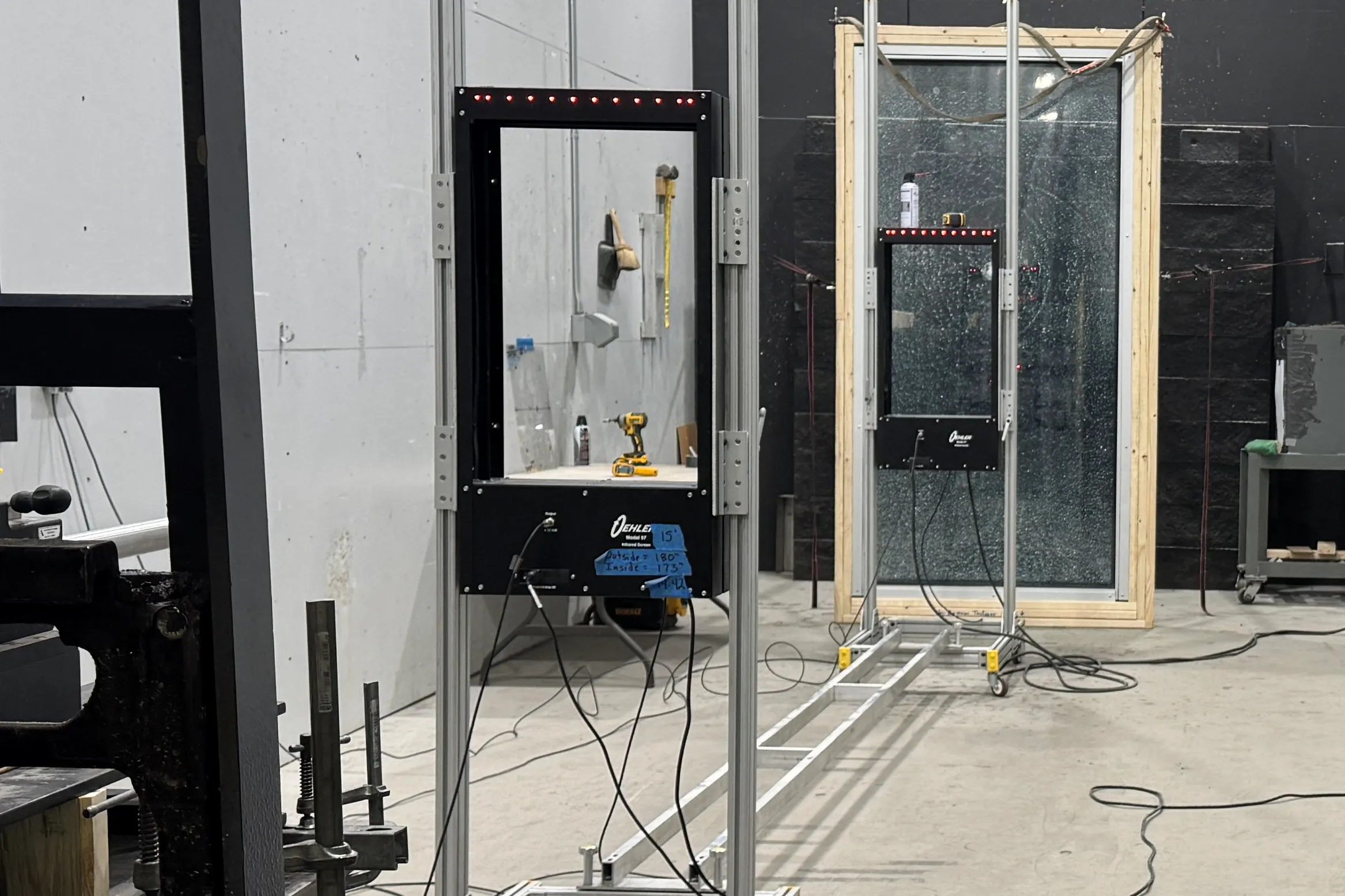 The forced entry test bay: two glazing frames on rails, chronograph screens, and a crazed laminated pane in a timber buck.